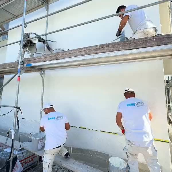 Drei Handwerker verputzen die Fassade, einer oben auf dem Gerüstbrett, zwei davor